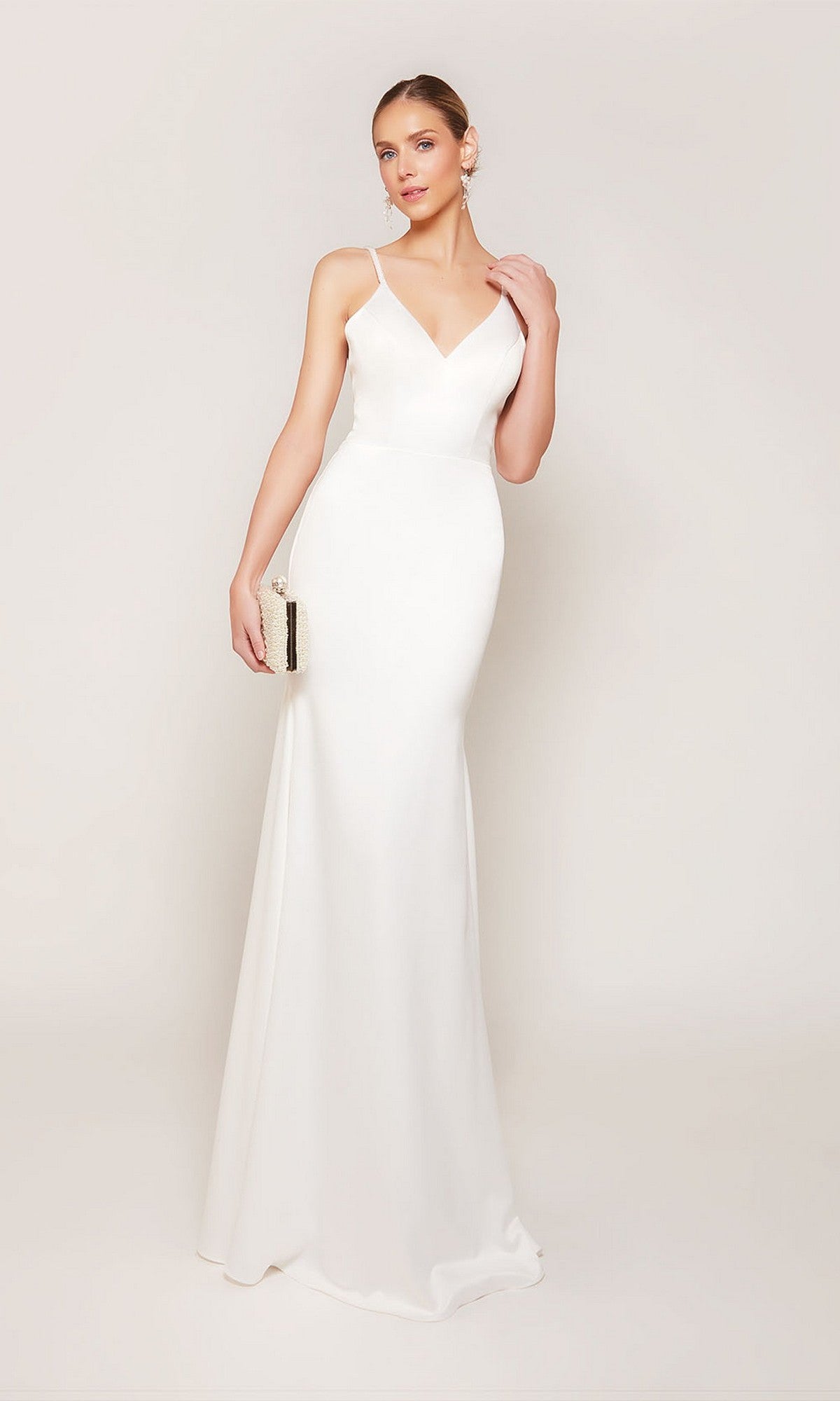 A woman in the Alyce 7090 Long Formal Dress, featuring a sleeveless, floor-length design in diamond white with thin straps, holds a small clutch against a plain light background.